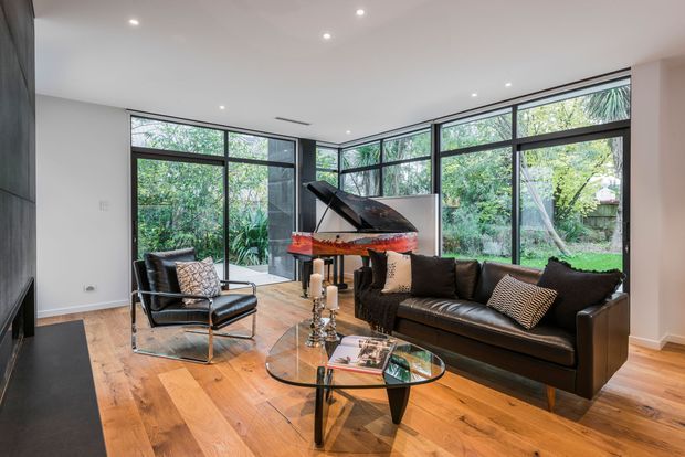 Bright, refined living room with full-height black-framed corner glazing overlooking lush greenery. The room features warm timber flooring, a black leather sofa and lounge chair, a glass coffee table, and a grand piano, with recessed ceiling lighting and a restrained contemporary fit-out.