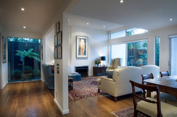 Open-plan living and dining area with warm timber flooring, cream walls, recessed lighting, and large windows and glazed doors connecting to a planted outdoor space. The living zone includes upholstered armchairs, a patterned rug, fireplace and artwork, while a timber dining table and chairs occupy the foreground. A glazed garden room or courtyard is visible to the left.