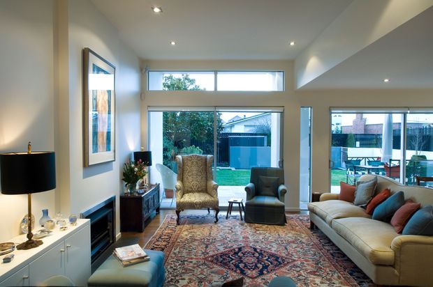 Bright contemporary living room with high ceilings, recessed downlights, large glazed openings to an outdoor area, a cream sofa, patterned armchair, dark fireplace surround, timber sideboard, and a large traditional patterned rug. The room has a warm, refined and inviting character.