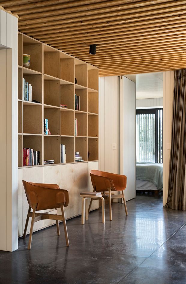 A warm, refined interior featuring a full-height open timber bookshelf with lower built-in cabinetry, two mid-century leather armchairs, and a small side table. The room has a dark polished concrete floor, vertical light wall panelling, and a striking exposed timber slat ceiling. A curtained opening leads toward a bedroom or sleeping area in the background.