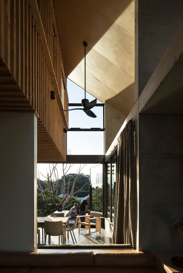 A tall, narrow interior view through a dramatic double-height glazed opening, showing a dining area beyond with a table and chairs, people, and garden views. The space features exposed concrete walls and beams, warm timber slatted wall and ceiling surfaces, a sculptural black ceiling fan, full-height curtains, and strong natural sunlight creating angular shadows. A partial sofa is visible in the foreground.