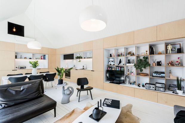 Bright open-plan living and dining room with a contemporary built-in timber wall and extensive white open shelving displaying books and decor. The room features a black leather sofa, white coffee table, dark dining chairs, pale flooring, sculptural pendant lights, and a high angled white ceiling.