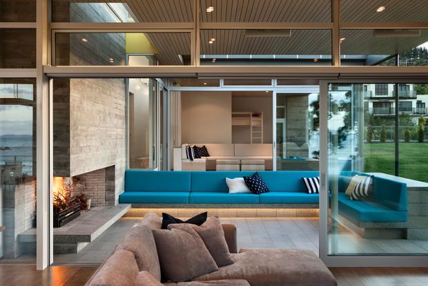 A contemporary living room with floor-to-ceiling glazed sliding doors, a turquoise built-in sectional sofa, a large brown lounge sofa in the foreground, and a substantial natural-stone fireplace wall. Timber-lined ceilings and extensive glazing create a bright connection to the landscaped exterior and water visible beyond.