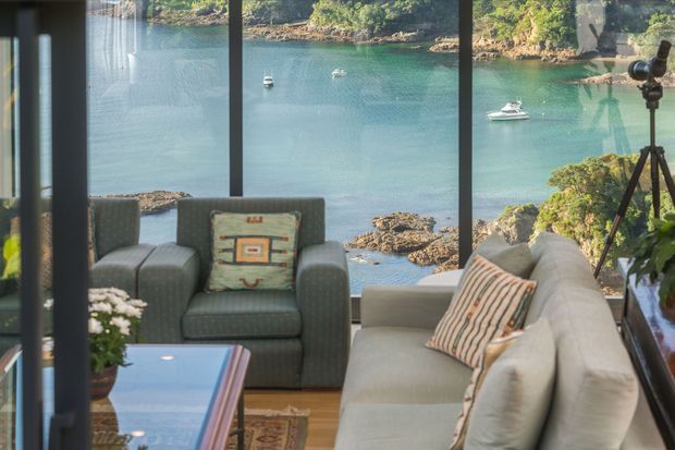 A furnished living room with a grey sofa and armchair arranged around a glass-topped coffee table. Full-height glazing provides a clear view over a sheltered turquoise-water inlet with rocky shoreline and small boats. A camera tripod and indoor plants are visible, while the room has a relaxed, coastal outlook.