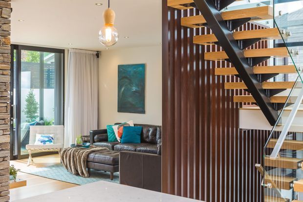 Open-plan living room with a contemporary floating staircase. The space features a dark leather sectional sofa, teal accents, a pale area rug, full-height glazing with curtains, a stone-clad column, and a sculptural pendant light. The staircase has timber treads, black steel stringers, glass balustrades, and a vertical dark timber slatted screen.