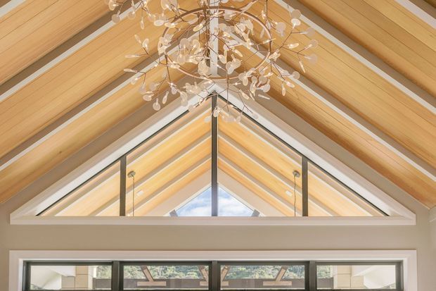 Upward view of a double-height vaulted interior with exposed light timber rafters and beams, clerestory glazing, and a dramatic branching chandelier. A high horizontal window band sits below the gable, bringing in natural light and reinforcing the airy architectural composition.
