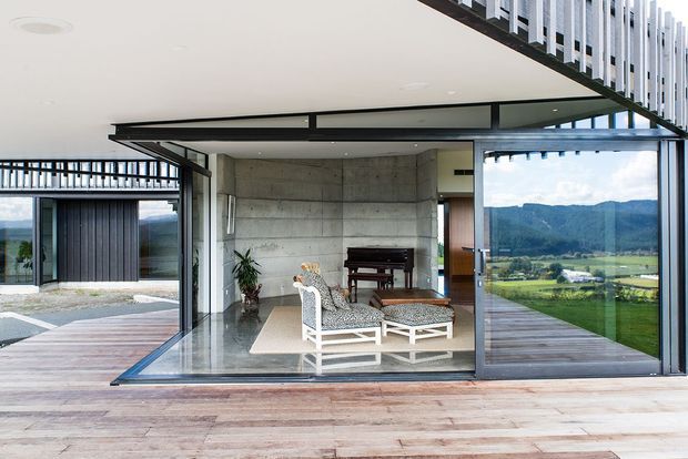 Open-plan living room with a piano, white lounge chairs, and a low coffee table, enclosed by extensive floor-to-ceiling glazing and large sliding glass doors. The space features a polished concrete floor, exposed concrete feature wall, timber decking extending through the room, and a dramatic projecting roof and upper-level screen. A green mountainous rural landscape is visible beyond the glazing.