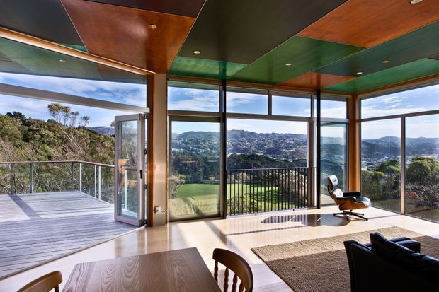 A sunlit contemporary living and dining space with floor-to-ceiling glazing opening onto a timber deck and expansive hillside views. The room features a richly toned timber dining table and chairs, dark leather lounge seating, a sculptural lounge chair, large woven rug, polished light flooring, and a dramatic multi-toned timber and green ceiling. Slim dark window frames and open glass doors create a strong connection to the outdoor terrace.