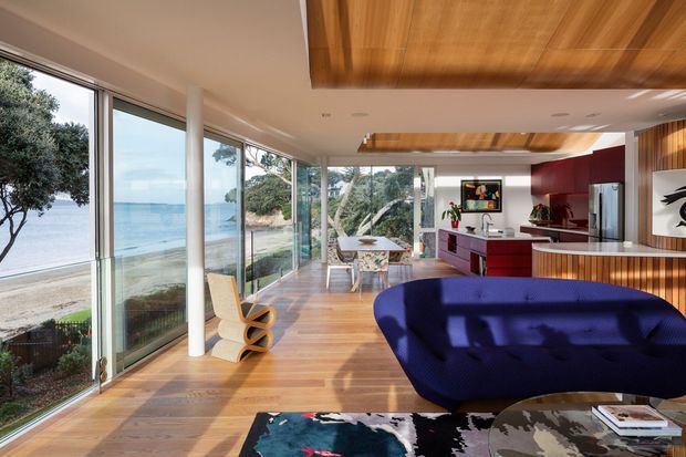 A bright, open-plan modern living and dining space overlooking a beach and water through extensive floor-to-ceiling glazing. The room features warm timber flooring, a richly timber-lined ceiling and built-in cabinetry, a deep blue sculptural sofa, a compact dining area, and a red-accented kitchen or bar zone. White structural columns and large sliding glass doors create a strong indoor-outdoor connection.