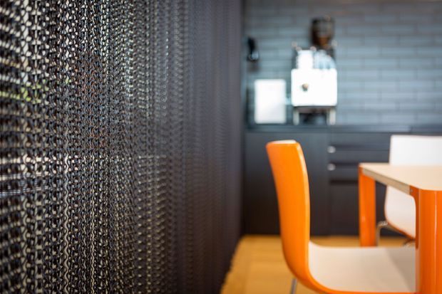 A close-up interior view of a compact contemporary kitchen or dining area. A black chain-link or mesh partition dominates the left side, while the background features dark cabinetry, a grey tiled splashback, and a white benchtop or appliance. Bright orange dining chairs and table legs provide a vivid accent against timber-toned flooring.