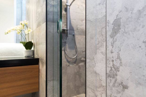 Close-up view of a contemporary bathroom featuring a frameless glass shower enclosure with a wall-mounted shower, large-format pale grey textured stone-look wall tiles, and a floating timber vanity with a dark benchtop. A white basin and small orchid arrangement are visible at the left.
