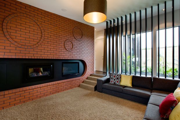 A contemporary living room with a dramatic exposed red-brick feature wall, integrated black fireplace and media joinery, large charcoal sectional sofa, and full-height windows screened by vertical black fins. A short set of steps leads toward a hallway, while a large cylindrical pendant provides warm illumination.