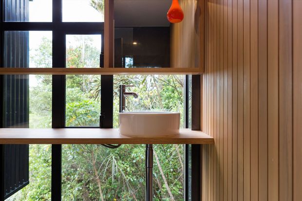 A compact, light-filled bathroom featuring a white vessel basin on a timber vanity shelf, with an exposed dark metal tap and plumbing. Black-framed windows overlook dense greenery, while vertical timber wall panelling and a warm orange pendant light add character.