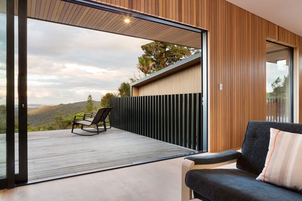 A contemporary indoor-outdoor living area opening onto a broad timber deck, with floor-to-ceiling sliding glazing, warm vertical timber wall cladding, a timber-lined ceiling, and a dark upholstered sofa. The deck overlooks a hilly natural landscape and connects to a covered outdoor zone with a black vertical-slat privacy screen.