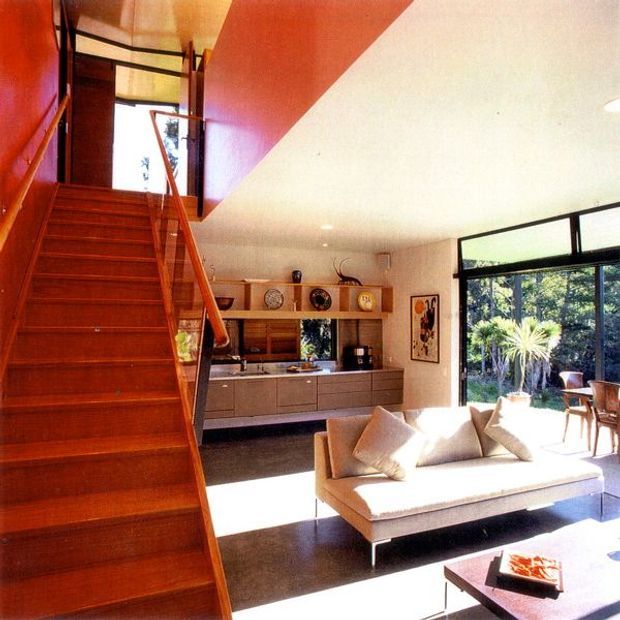 A bright, open-plan mid-century living space viewed from a timber staircase. The room features a warm timber stair and balustrade, a built-in timber and neutral-toned media/storage wall, a cream sofa, dark flooring, and large black-framed glazing opening toward a leafy garden. A strong red-orange accent wall and ceiling plane add graphic contrast, while the open stair and glazing create a spacious, connected interior.