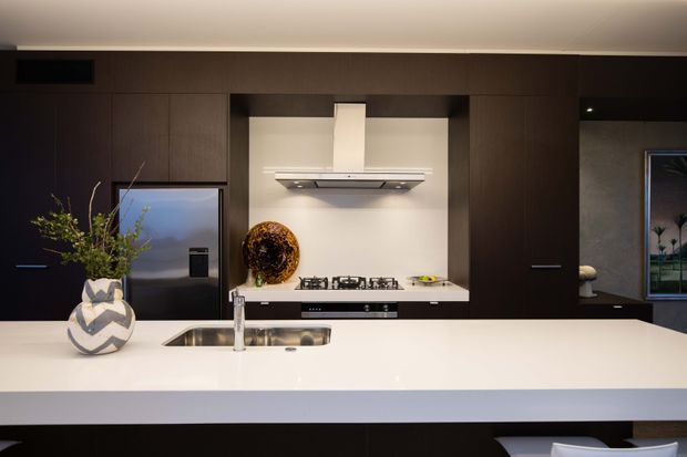 A contemporary kitchen viewed from the island, featuring dark timber cabinetry, a white stone island benchtop with integrated sink, a gas cooktop, white splashback, and concealed rangehood. Recessed display shelving and understated decorative objects add contrast, while a nearby opening provides a glimpse toward an adjacent space.