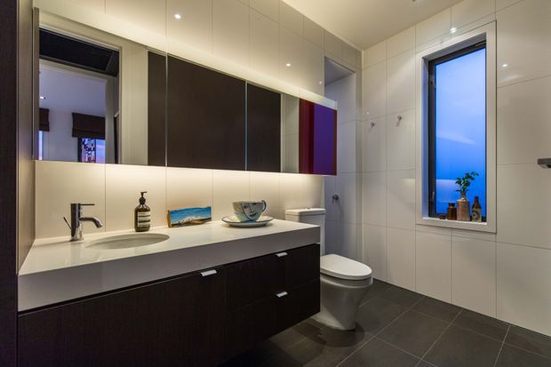 Contemporary bathroom with a wall-mounted dark timber vanity, integrated white basin and pale benchtop, large mirrored cabinetry, toilet, and full-height white wall tiles. Dark grey floor tiles create contrast, while a narrow window brings in blue evening light and a small potted plant.