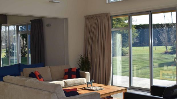 Bright contemporary living room with large floor-to-ceiling glazed sliding doors opening onto a manicured lawn. The room contains a large cream sectional sofa, dark leather seating, a low timber coffee table, neutral walls, and full-height taupe curtains.