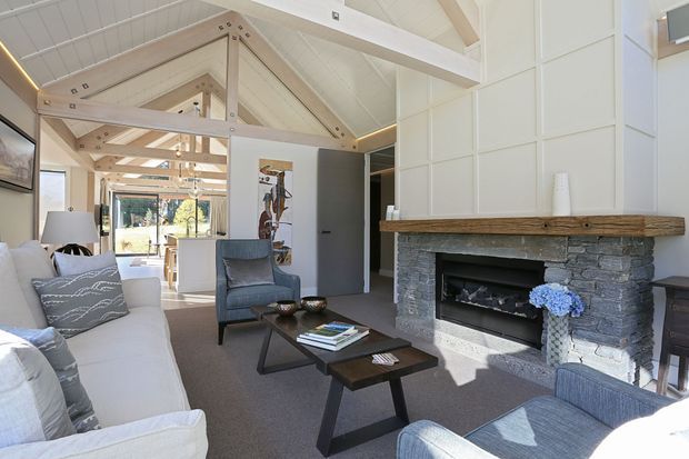 Bright, spacious living room with a vaulted white timber ceiling and exposed pale timber trusses, centered around a substantial stone fireplace with a natural timber mantel. The room features neutral upholstered seating, dark wood coffee tables, a grey carpet, white built-in wall panelling, and strong visual connection to a dining area and outdoors beyond.