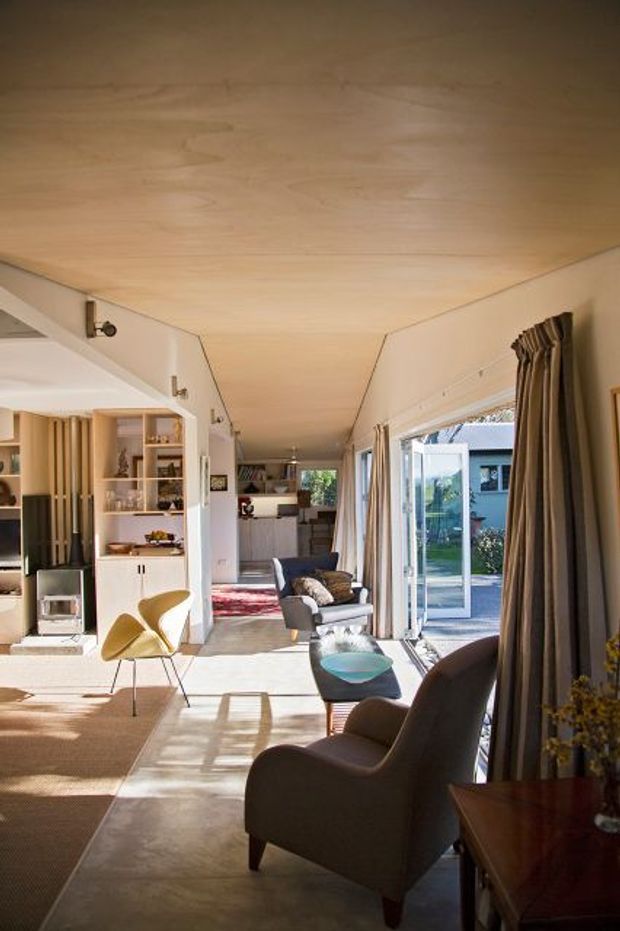 A long, sunlit open-plan living space with a warm timber-lined ceiling, polished concrete floor, extensive glazing and glazed doors opening to an outdoor garden area. The room contains mid-century-inspired lounge chairs, a low coffee table, built-in open shelving and cabinetry, and a rear kitchen or dining zone. Neutral white walls, timber joinery, and strong afternoon shadows create a warm, relaxed interior.