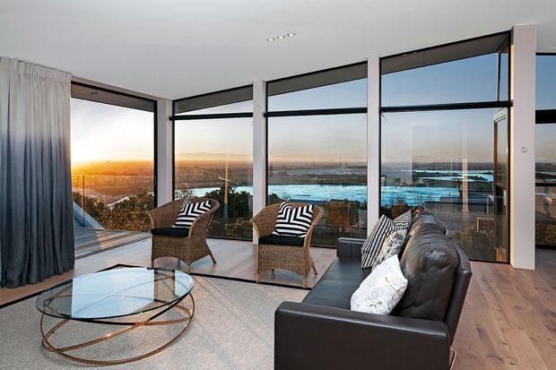 A refined contemporary living room with a dark leather sofa, woven lounge chairs, a round glass coffee table, and a large neutral rug. Floor-to-ceiling black-framed glazing wraps around the room, opening expansive views toward a turquoise waterfront and sunset. Timber flooring, white walls, recessed lighting, and full-height curtains complete the bright, elegant interior.