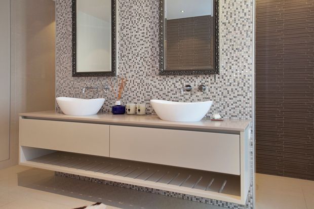 Contemporary double-vanity bathroom with two white vessel basins set on a long floating beige vanity. The wall behind is covered in small mosaic tiles, with two dark-framed mirrors, wall-mounted tapware, open lower shelving, and a dark timber-look tiled wall panel to the right.
