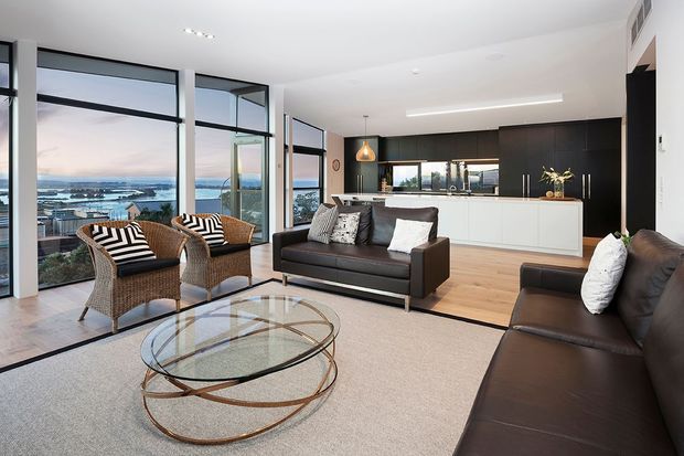 Bright open-plan living room with floor-to-ceiling glazing overlooking a waterfront or coastal harbour setting. The room features black leather sofas, woven armchairs with patterned cushions, a sculptural glass coffee table with a metal base, pale timber flooring, a light neutral carpet, and a sleek kitchen with white cabinetry and a dark island wall in the background.