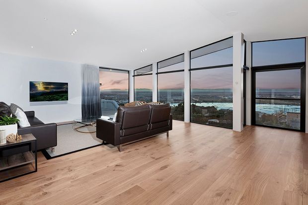 A bright contemporary living room with expansive floor-to-ceiling glazing overlooking a coastal urban landscape. The room features pale timber flooring, white walls and ceiling, dark leather sofas, a wall-mounted television, and streamlined black-framed windows and doors.