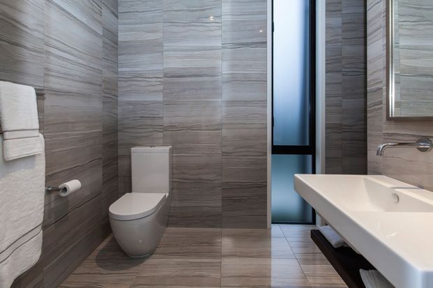Contemporary bathroom featuring full-height grey stone-look wall and floor tiles, a wall-mounted white basin with chrome tapware, a modern white toilet, folded white towels, and a dark-framed glazed door or window. The space is compact, polished, and evenly lit.
