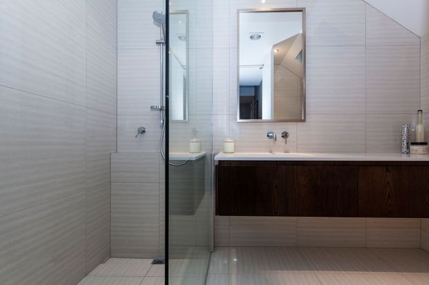 Contemporary bathroom with pale horizontally textured wall and floor tiles, a glass-enclosed walk-in shower, and a floating dark timber vanity with a light benchtop. A wall-mounted mirror and minimalist chrome fixtures complete the clean, understated design.