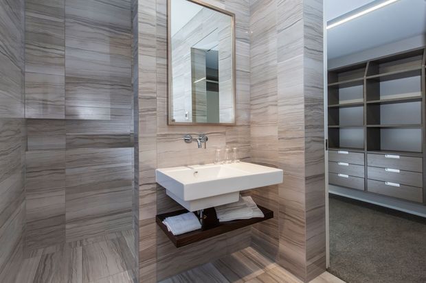 Contemporary bathroom vanity area clad in large-format grey-beige marble-look tiles, with a wall-mounted white basin, chrome wall-mounted tapware, framed mirror, and dark timber open shelving beneath. An open walk-in wardrobe with grey cabinetry and carpet is visible through the doorway on the right.