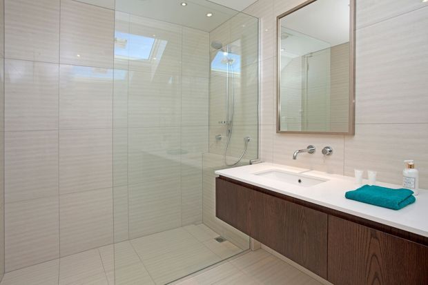 Bright contemporary bathroom with a large frameless glass walk-in shower, pale horizontally textured wall and floor tiles, a floating dark timber vanity with white integrated basin and benchtop, wall-mounted chrome tapware, and a large timber-framed mirror. A skylight and recessed ceiling lights provide abundant natural and artificial light.