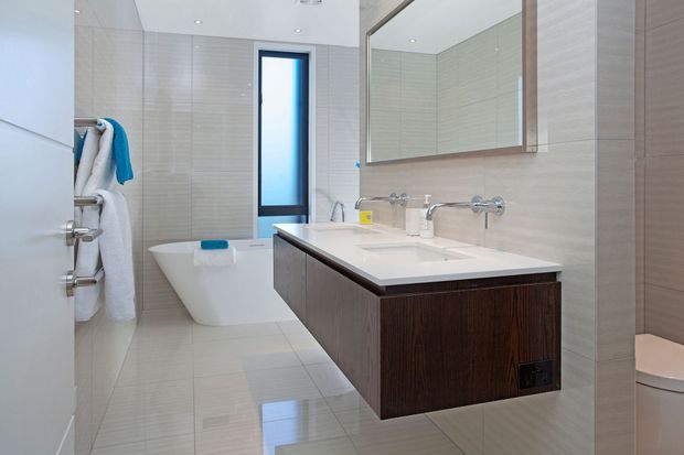 Bright contemporary bathroom with a freestanding white bathtub, wall-mounted double vanity, large framed mirror, pale tiled walls and floor, and dark timber-look floating cabinetry. A narrow vertical window provides natural light.