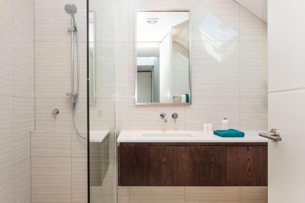 Bright contemporary bathroom featuring a frameless glass shower with built-in bench, pale large-format wall and floor tiles, a floating dark timber vanity with integrated basin, chrome tapware, and a wall-mounted mirror cabinet. The space is clean and compact with a neutral palette and strong natural light.