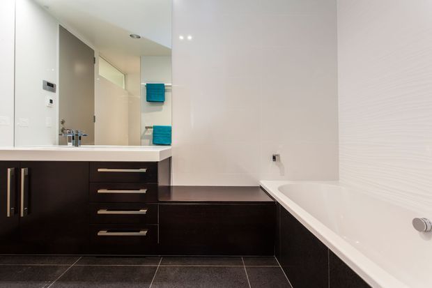 Modern bathroom with a large white bathtub, dark wood vanity and built-in cabinetry, pale tiled walls, large mirror, and dark stone-look floor tiles. Bright turquoise towels provide a small colour accent.