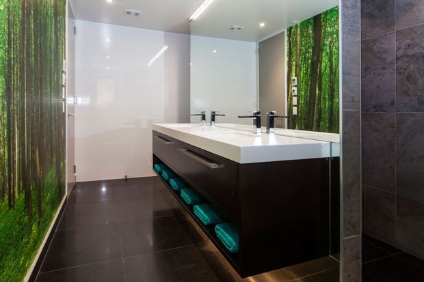 Contemporary bathroom with a large double vanity featuring dark built-in cabinetry, a thick white benchtop, twin basins and chrome tapware. Glossy white wall panels, dark floor tiles, dark wall tiles and a vivid forest-themed feature wall create a dramatic, polished interior.