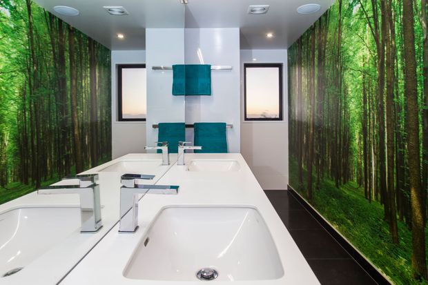 A bright contemporary bathroom with a large white double vanity and two integrated basins, freestanding-style inset bathtub, chrome geometric tapware, turquoise towels, dark floor tiles, and floor-to-ceiling photographic forest wall coverings on both sides. Small high-set windows and recessed ceiling lighting complete the room.