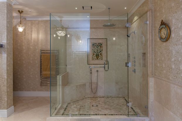 Luxurious traditional bathroom dominated by a large frameless glass walk-in shower with dual overhead and handheld fixtures. The room features ornate beige and cream textured wall finishes, decorative moulding, mosaic tile flooring, a heated towel rail, crystal-style wall lighting, and classic decorative accents including a wall clock and framed artwork.