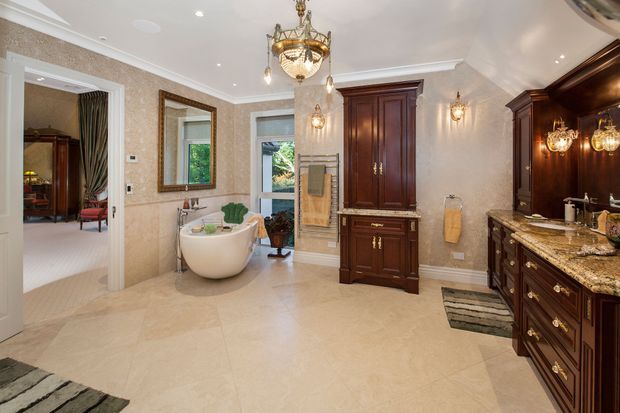 Luxurious traditional bathroom with a freestanding white soaking tub, large beige stone floor and wall tiles, dark stained timber vanity and cabinetry, ornate brass fixtures, decorative sconces, and a crystal chandelier. An open doorway reveals an adjoining carpeted bedroom or sitting area.