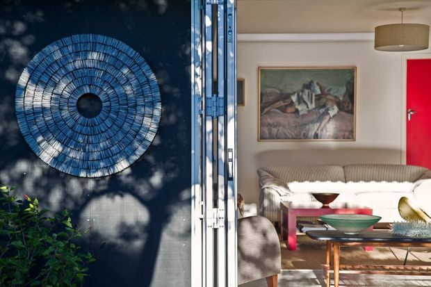 A split view from an open entry showing a dark blue exterior wall with a large circular woven wall feature and planting, alongside a bright living room. The interior contains a ribbed neutral sofa, sculptural coffee table, wood-framed artwork, pendant light, and a vivid red door, with strong patterned sunlight and shadows across the space.