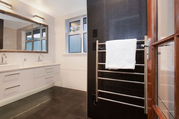 A contemporary bathroom with a double white vanity, two undermount basins, wall-mounted mirrors, dark large-format floor and feature wall tiles, frosted windows, and a chrome heated towel rail. Timber-framed glazing and warm integrated lighting add contrast to the monochromatic fit-out.