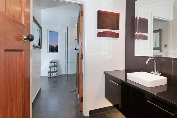 A contemporary bathroom viewed from the doorway, with a wall-mounted black vanity, white vessel basin, chrome tapware, large mirror, and dark tiled flooring and wall surfaces. A glass shower enclosure and hallway are visible beyond, with warm timber doors and red-toned artwork adding contrast.