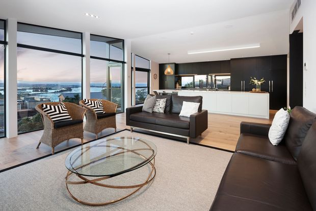 Bright contemporary open-plan living room with expansive floor-to-ceiling glazing overlooking a waterfront or harbour setting. The room features dark leather sofas, woven lounge chairs with patterned cushions, a round glass coffee table with a sculptural metal base, pale timber flooring, a large neutral rug, and a sleek open kitchen with white island cabinetry and dark tall cabinetry. Clean linear lighting and strong indoor-outdoor views create an airy, refined atmosphere.
