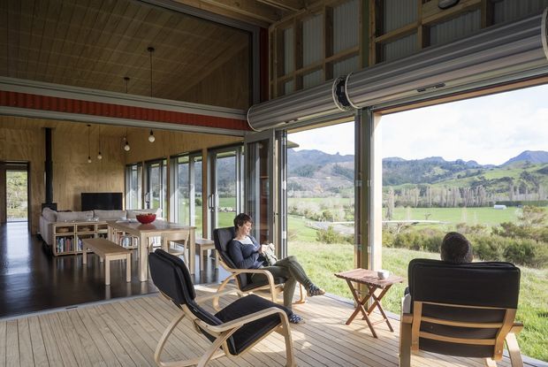 A bright, open-plan living and dining space with large retractable glazing opening onto a timber deck and expansive rural mountain views. The interior features a warm timber ceiling, dark flooring, exposed structural framing, pendant lighting, a dining table, and relaxed lounge chairs. A large horizontal roller blind or shading system is mounted above the glazing.