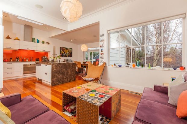 A bright open-plan living and kitchen area with polished timber flooring, white cabinetry, a dramatic dark stone island, and vivid red-orange illuminated backsplash. Large multi-pane windows bring in abundant natural light, while purple sofas, a sculptural leather lounge chair, colourful pendant lights, and a patterned coffee table add eclectic character. The room has detailed traditional-style ceiling mouldings and opens toward a dining area.