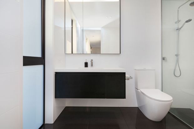 Bright contemporary bathroom with a wall-mounted dark vanity, white basin, frameless mirror, wall-hung toilet, and glass-enclosed shower. Large dark floor tiles contrast with the predominantly white walls and sanitaryware, while black-framed glazing adds a strong linear detail.