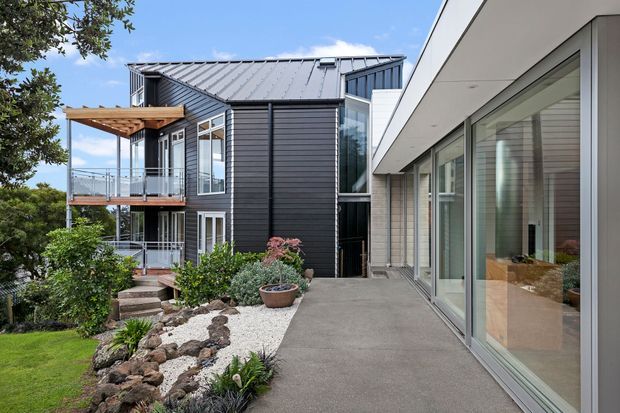 Exterior view of a contemporary multi-level residential building on a landscaped sloping site. The composition shows dark horizontal cladding, a pitched metal roof, extensive glazing, balconies with timber elements, and a light-coloured rendered or panelled wing connected by a broad paved terrace and garden landscaping.