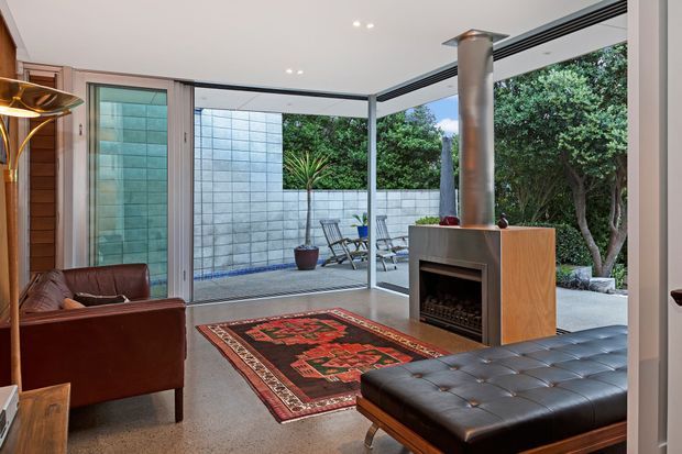 A refined indoor-outdoor living room with full-height glazed sliding doors opening to a paved courtyard. The room features a freestanding fireplace clad in warm timber, a polished concrete floor, leather seating, a patterned area rug, timber wall panelling, and a covered outdoor terrace with a concrete block privacy wall and planting. The space has strong mid-century modern character with contemporary detailing.
