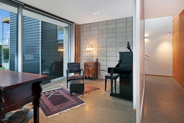 A refined modern interior combining a piano sitting area with a dining space and hallway. The room features polished concrete flooring, a grey concrete-block feature wall, expansive glazing with dark horizontal exterior cladding visible beyond, warm timber wall panelling in the corridor, and layered rugs and leather seating.