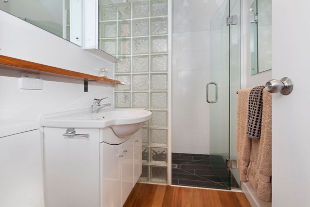 Compact bright bathroom with a white vanity and basin, wall-mounted mirror cabinetry, a timber shelf, glass-block partition, and a frameless glass shower enclosure. The space combines white tiled walls with dark floor tiles and warm timber flooring, creating a clean, practical fit-out.